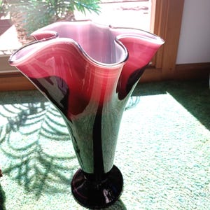 Op de afbeelding: Een decoratieve glazen vaas met een gegolfde rand. De vaas heeft een kleurengradiënt, van een diepe bordeauxrode basis tot een lichter roze aan de bovenkant. De vaas heeft een uitlopende vorm en een ronde basis.