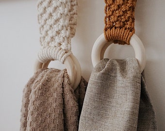 Puede incluir: Dos anillos de madera con bucles colgantes de macramé, uno con una toalla beige y el otro con una toalla marrón claro.