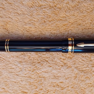 Puede incluir: Una pluma estilogr&aacute;fica negra con detalles dorados y un clip dorado. La pluma est&aacute; sobre una superficie peluda de color marr&oacute;n claro.