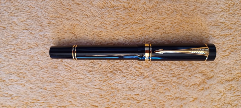 Puede incluir: Una pluma estilogr&aacute;fica negra con detalles dorados y un clip dorado. La pluma est&aacute; sobre una superficie peluda de color marr&oacute;n claro.