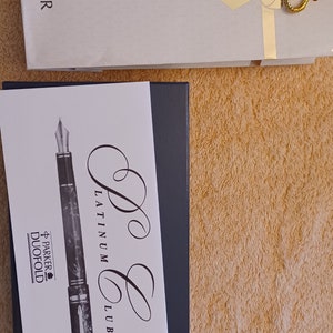 Puede incluir: Una imagen en blanco y negro de una pluma estilogr&aacute;fica Parker Duofold con un dise&ntilde;o de m&aacute;rmol. La pluma est&aacute; en una caja con las palabras "Parker Platinum Club" escritas en ella.
