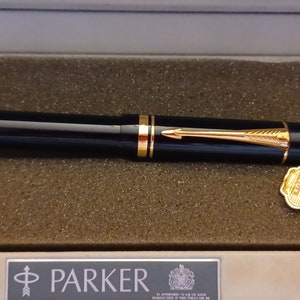 Puede incluir: Una pluma estilogr&aacute;fica Parker negra con detalles dorados y un capuch&oacute;n dorado en una caja negra y blanca. La pluma est&aacute; en una caja con el logotipo de Parker y un sello dorado.