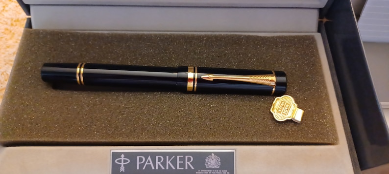 Puede incluir: Una pluma estilogr&aacute;fica Parker negra con detalles dorados y un capuch&oacute;n dorado en una caja negra y blanca. La pluma est&aacute; en una caja con el logotipo de Parker y un sello dorado.