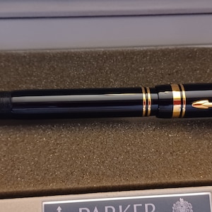 Puede incluir: Una pluma estilogr&aacute;fica Parker negra y dorada en una caja negra con ribete dorado. La pluma tiene una plumilla dorada y una banda dorada alrededor del cuerpo. La caja tiene una etiqueta dorada con el logotipo de Parker.