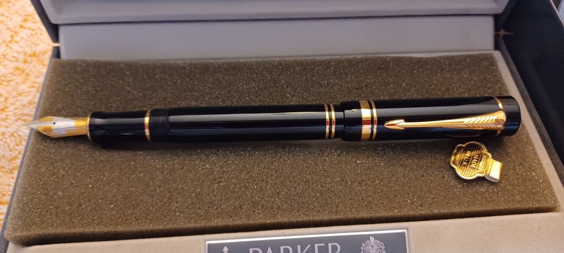Puede incluir: Una pluma estilogr&aacute;fica Parker negra y dorada en una caja negra con ribete dorado. La pluma tiene una plumilla dorada y una banda dorada alrededor del cuerpo. La caja tiene una etiqueta dorada con el logotipo de Parker.
