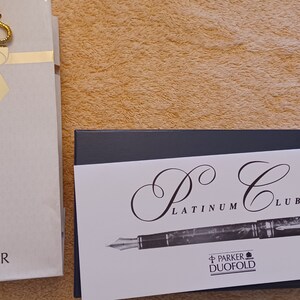 Puede incluir: Un bol&iacute;grafo fuente Parker Duofold Platinum Club en una caja negra con una tarjeta blanca que dice "P.C. Platinum Club" y "Parker Duofold".