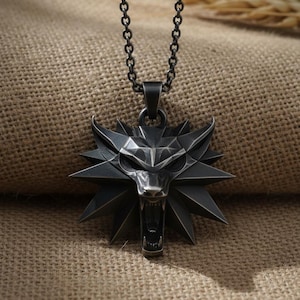 May include: A silver-colored pendant necklace featuring a stylized wolf head with an open mouth and sharp teeth. The pendant is attached to a chain necklace. The background is a textured, tan fabric.