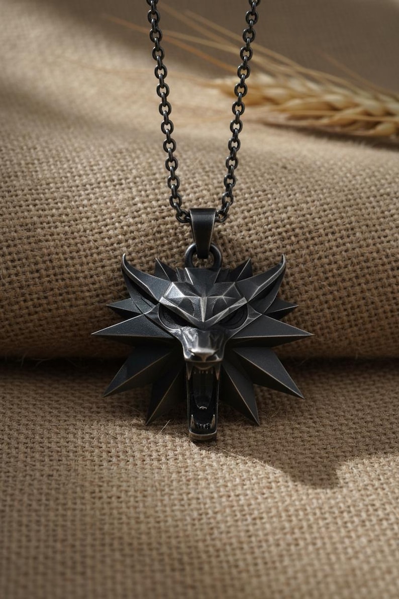 May include: A silver-colored pendant necklace featuring a stylized wolf head with an open mouth and sharp teeth. The pendant is attached to a chain necklace. The background is a textured, tan fabric.