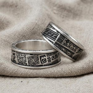 May include: Two silver-toned metal rings featuring intricate, dark, engraved geometric patterns covering the entire outer surface. The bands have a wide, flat profile with detailed, repeating symbols and lines etched into the metallic finish.