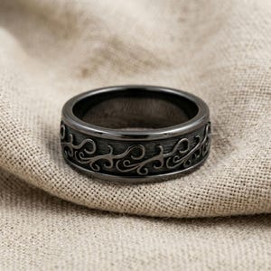 May include: A dark gunmetal gray ring featuring a continuous, raised, swirling vine-like pattern engraved around the outer band. The metal has a polished finish on the edges and a brushed, textured appearance within the recessed scrollwork design.