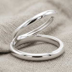 Puede incluir: Un anillo de tono plateado pulido con un diseño de doble banda. El anillo consta de dos bucles de metal lisos y redondeados que están unidos por un lado, creando una apariencia abierta y superpuesta con un acabado reflectante de alto brillo.