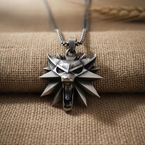 May include: A silver-colored pendant necklace featuring a stylized wolf head design. The pendant has a geometric, angular appearance with an open mouth and sharp teeth. The necklace chain is a fine, linked style.