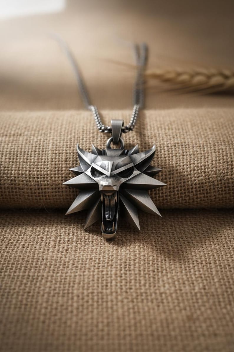 May include: A silver-colored pendant necklace featuring a stylized wolf head design. The pendant has a geometric, angular appearance with an open mouth and sharp teeth. The necklace chain is a fine, linked style.