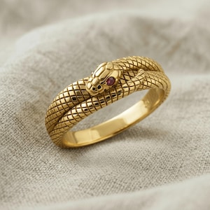 May include: A gold ring shaped like a coiled snake with a textured scale pattern covering the entire band. The snake head features a small, round, deep red gemstone set as an eye, creating a detailed and realistic appearance for this piece of jewelry.