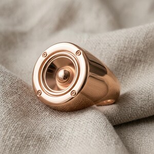 A close-up image of a gold-colored ring with a speaker design, resting on a beige fabric surface.