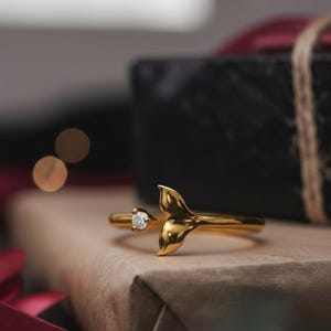 May include: A gold-colored ring with a mermaid tail design and a small, clear gemstone. The ring is open, with the tail on one end and the gemstone on the other. The ring is resting on a wrapped gift box.