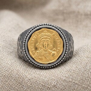 May include: A ring featuring a central gold-toned coin with an embossed portrait of a figure wearing a crown and holding an orb. The coin is set within a dark, oxidised silver band that has a textured, hammered finish and a beaded border.