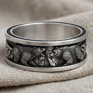 May include: A silver-toned metal ring featuring a detailed, embossed relief of two bears facing each other. The band has a smooth, polished top and bottom edge, with a darkened, recessed background that highlights the textured fur of the bears.