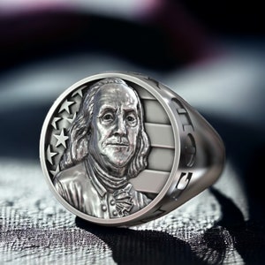 Könnte beinhalten: Ein silberner Ring mit einer runden Fläche, die ein Porträt von Benjamin Franklin und eine stilisierte amerikanische Flagge zeigt. Die Ringschiene ist mit dem Wort "Liberty" beschriftet.
