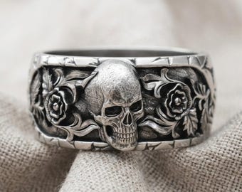 Könnte beinhalten: Ein breiter Ring aus silberfarbenem Metall mit einem zentralen, geprägten Totenkopfmotiv, das von detaillierten Blumen- und Blattverzierungen umgeben ist. Das Band hat eine texturierte, oxidierte Oberfläche, die das Relief hervorhebt.