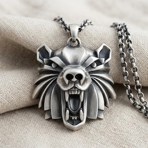 May include: A silver-toned metal pendant shaped like a snarling bear head with visible teeth and fur detailing, suspended from a matching silver-toned metal chain link necklace. The pendant features a dark, oxidized finish in the recessed areas.