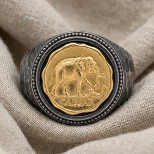 May include: A ring featuring a dark, hammered metal band with a circular bezel. The center displays a gold-toned coin-like inlay depicting an elephant and a snake, with the word CAESAR inscribed in capital letters at the bottom of the coin design.