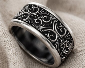 May include: A wide silver ring featuring a dark, recessed background with intricate, raised scrollwork and swirling floral patterns. The metal has a polished finish on the edges, contrasting with the blackened, detailed texture of the band.