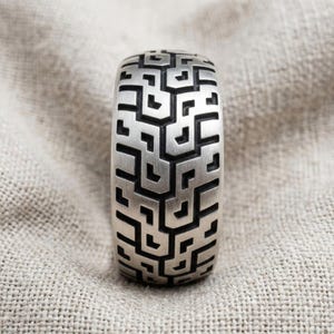 May include: A wide silver band ring featuring a continuous, recessed geometric maze pattern. The metal surface has a brushed, matte finish, while the deep, intricate grooves of the labyrinth design are darkened to create a high-contrast visual effect.