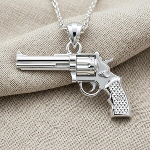 Könnte beinhalten: Ein silberfarbener Revolver-Anhänger aus Metall mit detailliertem Lauf, Trommel und strukturiertem Griff. Der Anhänger hängt an einer passenden silberfarbenen Metallkette und zeichnet sich durch eine polierte Oberfläche und komplexe mechanische Details aus.
