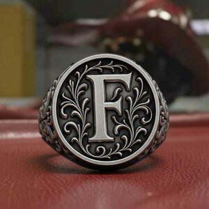 Könnte beinhalten: Ein silberner Siegelring mit dem Buchstaben "F" in der Mitte, umgeben von einem verzierten floralen Design. Der Ring liegt auf einer roten Oberfläche, mit unscharfem Hintergrund.