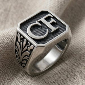 May include: A silver-toned signet ring featuring a square face with the letters CF engraved in a bold serif font against a dark, textured background. The sides of the ring are decorated with an embossed floral pattern with smooth, polished silver details.