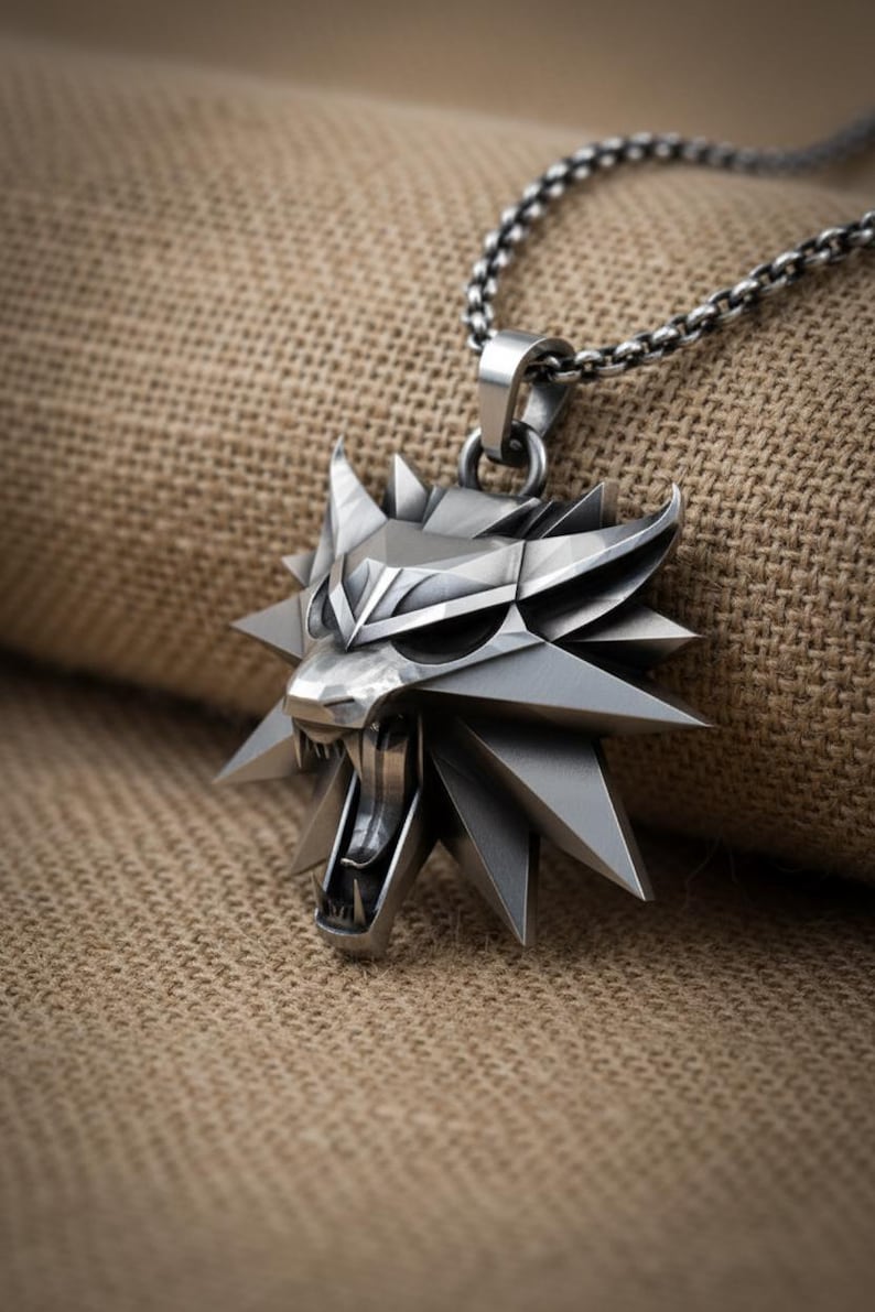 May include: A silver-colored pendant necklace featuring a stylized wolf head with sharp angles and open mouth. The pendant is attached to a chain necklace. The background is a textured, tan fabric.