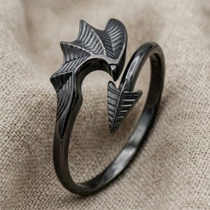 May include: A black metal cuff bracelet with an open design. The bracelet features a stylized wing and arrow design. The wing has detailed feather-like etching, and the arrow has a textured surface. The bracelet is set against a neutral background.