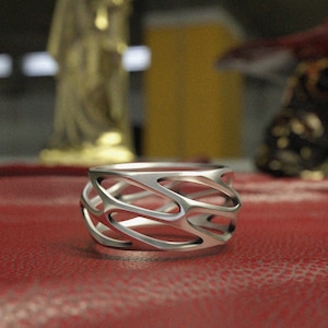 May include: A silver ring with an open, interwoven design. The ring has a wide band with a series of curved, elongated openings. The ring is displayed on a red surface, with blurred background elements including a gold statue.