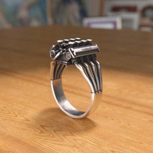 Puede incluir: Un anillo de plata con un diseño de motor detallado. El anillo presenta un pequeño motor con tubos de escape que forman la banda del anillo.