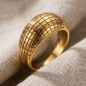 Puede incluir: Un anillo de cúpula en tono dorado con acabado pulido y un patrón de cuadrícula repetitivo de hendiduras cuadradas en toda la superficie exterior. La banda tiene un perfil ancho y redondeado con un interior liso y textura metálica.