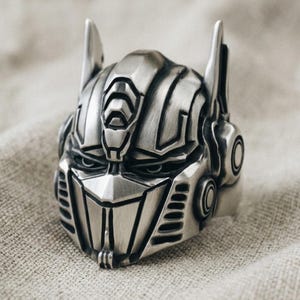 Könnte beinhalten: Ein silberfarbener Metallring in Form eines Roboterhelms mit spitzer Frontplatte, kantiger Stirnpartie und zwei vertikalen Antennen an der Oberseite. Die Oberfläche ist gebürstet und weist tiefe Rillen auf, die die mechanischen Details betonen.