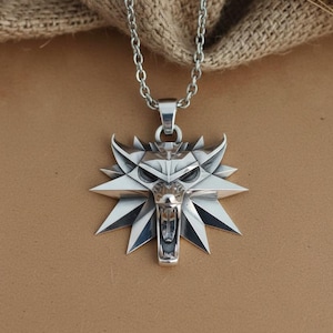May include: A silver-colored pendant necklace featuring a wolf head design. The pendant has a detailed, angular wolf head with an open mouth and sharp features. The necklace chain is also silver and hangs against a neutral background.