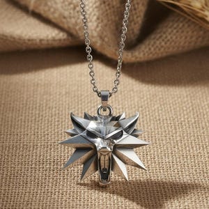 May include: Silver-colored pendant necklace featuring a wolf head emblem with a geometric design. The pendant is suspended from a delicate chain. The necklace is displayed against a neutral, textured background.