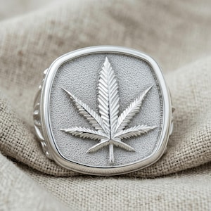 May include: A silver-toned ring featuring a square face with rounded corners. The center of the face displays a raised, detailed cannabis leaf design set against a textured, matte silver background. The band has a polished, smooth finish.