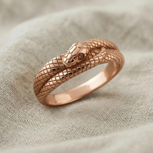 Op de afbeelding: Een roségouden ring in de vorm van een kronkelende slang met een gestructureerd, schubachtig patroon over de gehele band. De slangenkop is voorzien van een kleine, ronde, donkerrode edelsteen als oog, waarbij het lichaam zich rond de ring vormt.