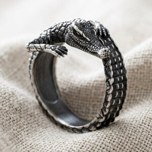 Könnte beinhalten: Ein silberfarbener Metallring in Form eines Krokodils mit strukturierter, schuppiger Oberfläche. Das Design zeigt den Kopf und die Vorderbeine des Krokodils, die über den Ring ragen und einen offenen Wickelstil mit dunklen, oxidierten Details bilden.