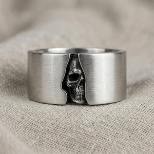 Può includere: Un anello color argento con un design a teschio. L'anello ha una fascia larga in metallo spazzolato. Il teschio è intagliato al centro dell'anello, creando un'estetica audace e gotica.