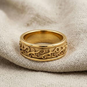 Può includere: Un anello dorato con un intricato motivo a spirale. L'anello ha una fascia larga e una superficie strutturata, che mostra un design dettagliato di linee e curve fluide. L'anello è presentato su un tessuto di colore neutro.