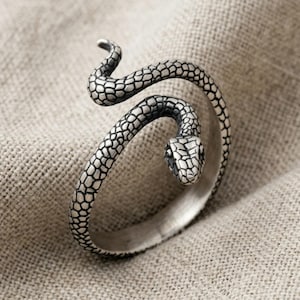 Op de afbeelding: Een zilverkleurige ring in de vorm van een opgerolde slang met een gestructureerd, schubachtig patroon over het gehele oppervlak. De slangenkop bevindt zich aan één uiteinde van de open ring en is voorzien van gedetailleerde markeringen.