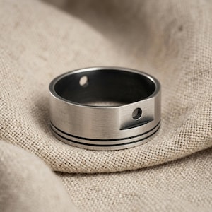 Könnte beinhalten: Ein breiter Ring aus gebürstetem, silberfarbenem Metall mit flachem Banddesign und zwei dünnen, horizontalen schwarzen Gravurlinien, die den unteren Teil umkreisen. Der Ring hat einen erhöhten rechteckigen Bereich mit einem kleinen Loch.