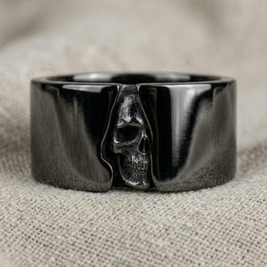May include: A wide, polished black ring featuring a detailed, three-dimensional skull relief centered within a vertical gap. The band has a smooth, reflective finish that contrasts with the textured, weathered appearance of the skull design.