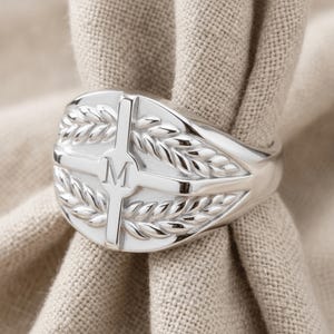 May include: A polished silver-tone signet ring featuring an oval face with a central vertical bar engraved with the letter M. The design includes symmetrical laurel leaf motifs flanking the center bar, creating a detailed relief pattern.