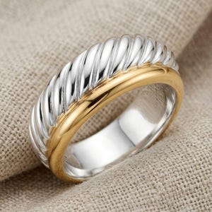 May include: A two-tone band ring featuring a polished silver-coloured section with a prominent twisted rope texture and a smooth, solid gold-coloured section. The ring has a wide profile and a reflective metallic finish on both the twisted and smooth parts.
