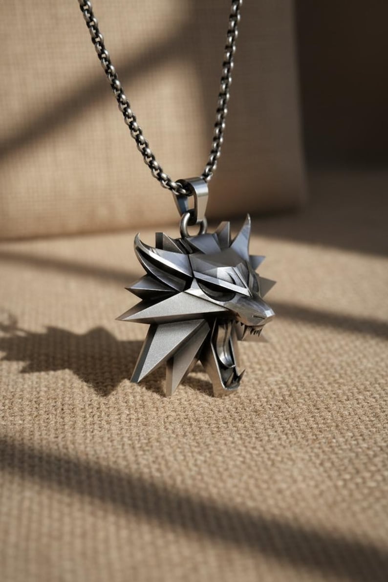 May include: Silver-toned pendant necklace featuring a stylized wolf head with an open mouth and sharp teeth. The pendant is suspended from a matching chain. The design has a geometric, angular aesthetic, and the necklace is likely made of metal.